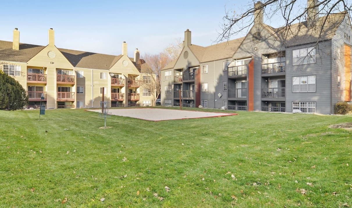Three-story Habitat at Fort Collins apartment buildings with balconies surround a well-kept lawn and a sand volleyball court under a clear sky. Trees and shrubs are visible around the grassy area in Fort Collins.