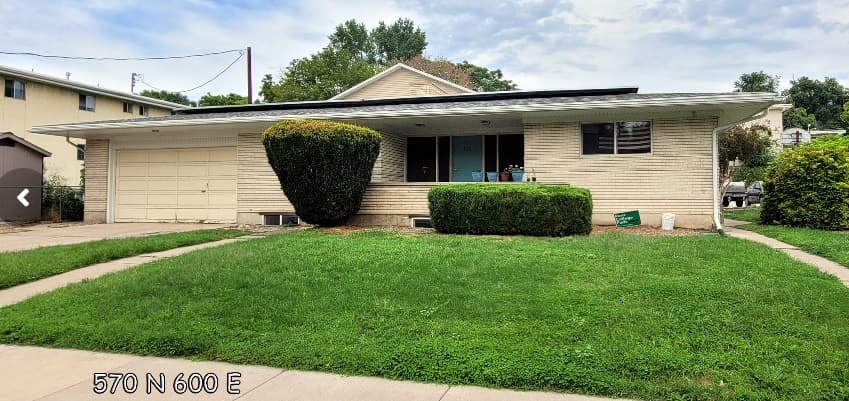 Charming single-story beige home in Autumn Hills Cove, Logan, featuring a wide front lawn, two trimmed bushes, an attached two-car garage, a covered porch with light blue doors, and a sidewalk along the edge of the grass.