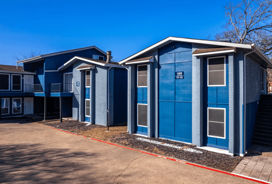 Treehouse Apartments features two modern blue buildings with white trim, flat roofs, and multiple windows under a clear blue sky. A paved driveway and minimal landscaping complete the inviting exterior.