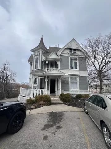 A large, gray-and-white Victorian-style house with a pointed turret and a covered porch, surrounded by leafless trees and parked cars, under a cloudy sky.