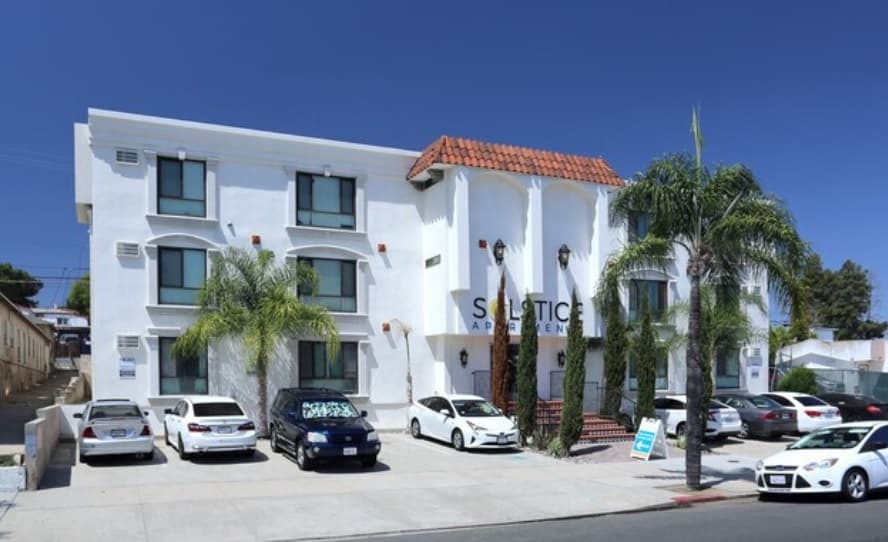 A white three-story apartment building with a red-tiled roof and arched windows, featuring a prominent “Solstice Apartments” sign. Several cars are parked in front, while palm trees and bright sunshine evoke the warmth of Solstice days.