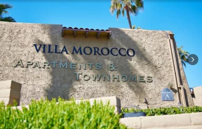 A beige stucco building displays a “VILLA MOROCCO APARTMENTS & TOWNHOMES” sign in blue letters, with palm trees and a bright blue sky, capturing the serene charm of Villa Morocco.