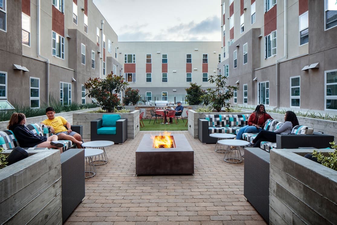 Residents at M at College San Diego sit and relax on cushioned outdoor furniture around a rectangular fire pit in a modern apartment courtyard, surrounded by greenery, tables, and multi-story buildings in the background.