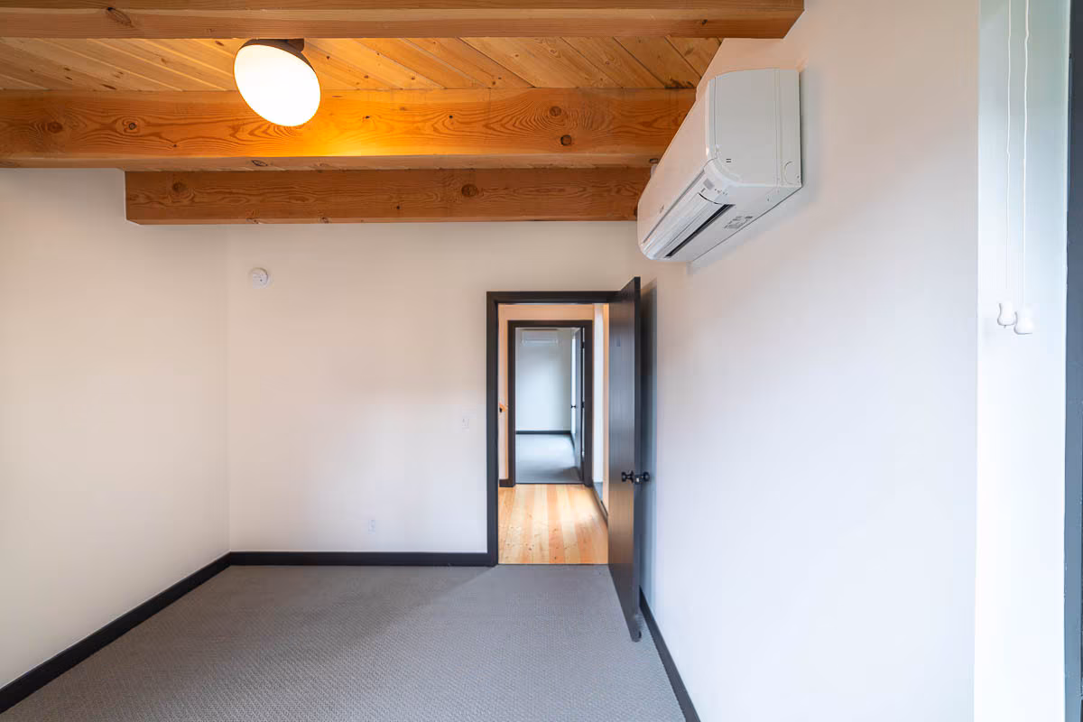 A bright, empty room with beige walls, a gray carpet, exposed wooden beams, and an air conditioning unit—perfect for joining the Village Coliving community. An open door leads to a hallway with warm wooden floors.