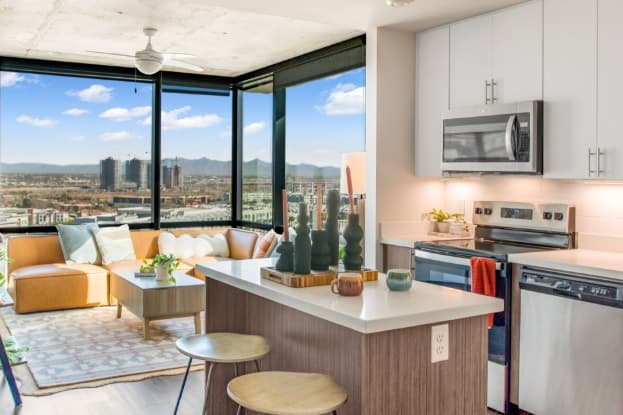 Modern apartment with floor-to-ceiling windows, a city skyline view, cozy living area with a tan sofa, and sleek kitchen—perfect for ASU Housing or anyone seeking stylish University Housing near campus.